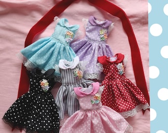 May include: A collection of miniature doll dresses in various colors, including blue, purple, red, black, and pink. Each dress features a polka dot or striped pattern, lace trim, and a floral embellishment. A red velvet ribbon is at the top.