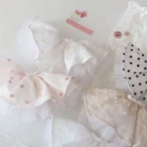Op de afbeelding: Een set van vier witte en roze babyjurkjes voor meisjes met verschillende patronen, waaronder hartjes, stippen en kant. De jurkjes zijn gemaakt van zachte, delicate stof en zijn perfect voor speciale gelegenheden.
