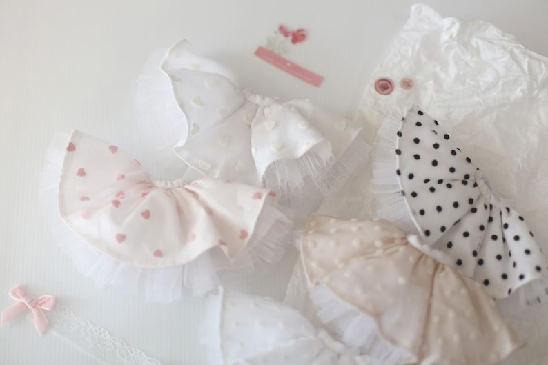 Op de afbeelding: Een set van vier witte en roze babyjurkjes voor meisjes met verschillende patronen, waaronder hartjes, stippen en kant. De jurkjes zijn gemaakt van zachte, delicate stof en zijn perfect voor speciale gelegenheden.