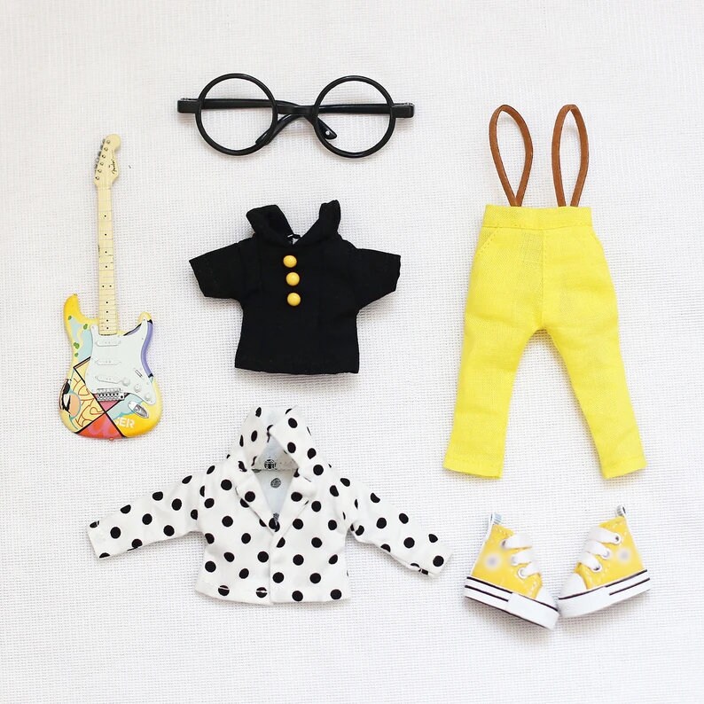 May include: A set of doll clothes including a black short-sleeved shirt, yellow pants with suspenders, a white polka dot jacket, and yellow sneakers.
