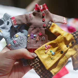 May include: Four miniature doll clothes with embroidered floral designs. The clothes are in shades of brown, gray, yellow, and red.