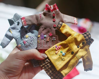 May include: Four miniature doll clothes with embroidered floral designs. The clothes are in shades of brown, gray, yellow, and red.