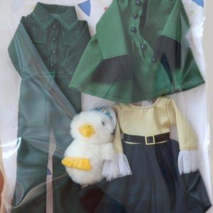 Puede incluir: Un conjunto de ropa de muñeca en verde y negro, que incluye un vestido de manga larga, una chaqueta, un pantalón y una falda. También se incluye un pato de peluche blanco con un pico amarillo.