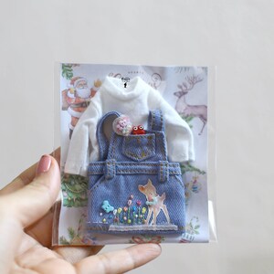 May include: A blue denim overall dress with a white long-sleeved shirt, a small red frog, and a flower embellishment. The dress has a deer embroidered on the front with flowers and butterflies.