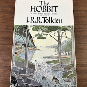May include: A vintage copy of "The Hobbit" by J.R.R. Tolkien, a two-sided jigsaw puzzle. The cover features a landscape illustration with a river, trees, and a small village. The title and author's name are printed in black.