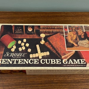May include: A vintage Scrabble Sentence Cube Game box with a brown wooden background. The box features a picture of two people playing the game with letter cubes. The box is labeled "Scrabble Sentence Cube Game" and "1971 Selchow & Righter Co."