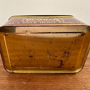 May include: A vintage Crayola crayon tin, rectangular with a trapezoidal shape, in shades of brown and gold. The top features the Crayola logo and the text "USE A GOLD MEDAL PRODUCT". The tin shows signs of age and wear.