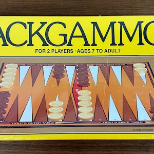 May include: Backgammon game box with a yellow background and the game's name in large black letters. The box is labeled "FOR 2 PLAYERS AGES 7 TO ADULT." The game board features brown, white, and orange triangles, with dice and checkers.