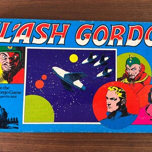 May include: A Flash Gordon board game box cover featuring a blue background with a spaceship, planets, and characters from the comic book series. The text "FLASH GORDON" is in large, white letters at the top of the box. The text "Adventure on the Moons of Mongo Game" is in smaller, white letters below the title. The box is designed for 2 to 4 players and is suitable for ages 6 to adult.