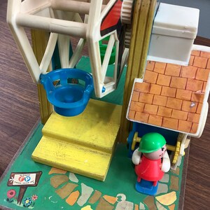 K&ouml;nnte beinhalten: Vintage Fisher-Price Little People Spielset mit einem Riesenrad, einer gelben Plattform, einem blau-wei&szlig;en Riesenrad, einem Geb&auml;ude mit gr&uuml;nem und rotem Dach und einer roten und gr&uuml;nen Little People Figur. Die Basis des Spielsets ist gr&uuml;n mit einem braunen Weg und einem Schild mit der Aufschrift "Ein Original Fisher-Price Spielzeug".