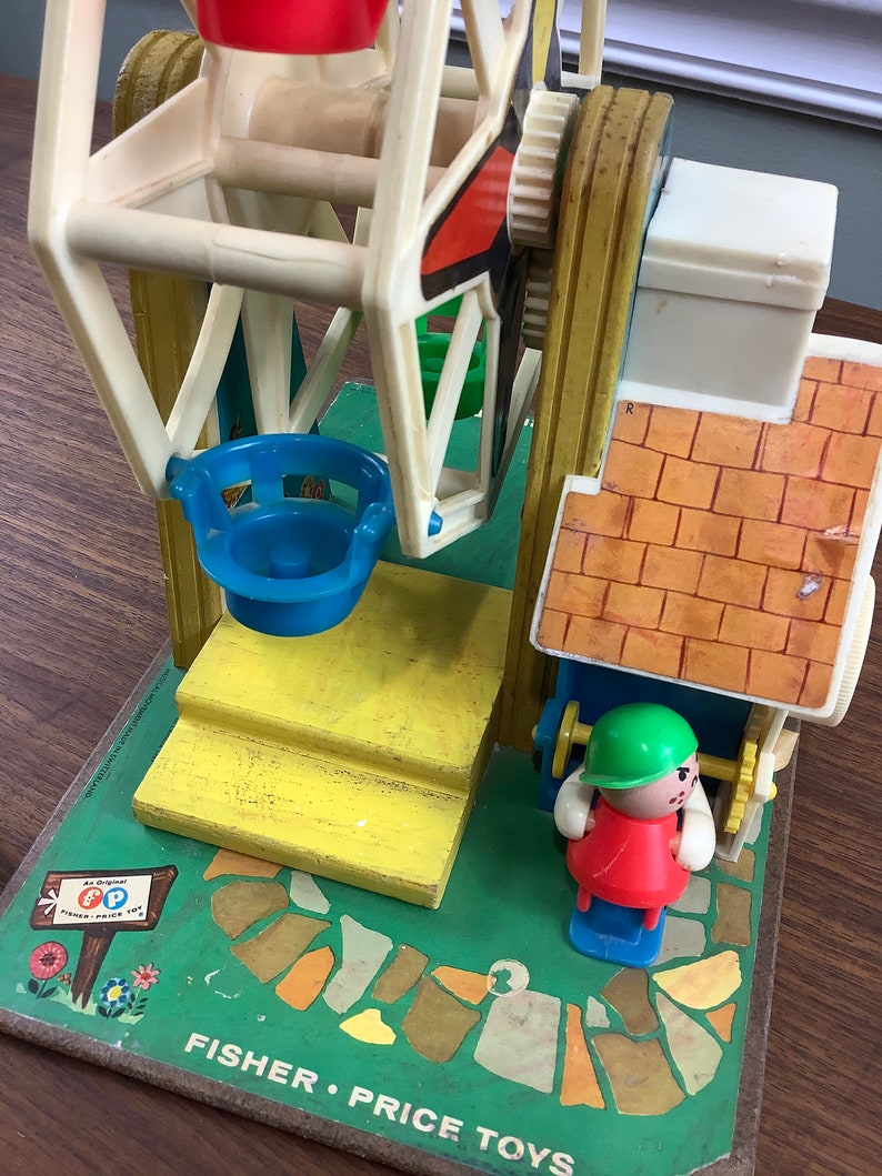 K&ouml;nnte beinhalten: Vintage Fisher-Price Little People Spielset mit einem Riesenrad, einer gelben Plattform, einem blau-wei&szlig;en Riesenrad, einem Geb&auml;ude mit gr&uuml;nem und rotem Dach und einer roten und gr&uuml;nen Little People Figur. Die Basis des Spielsets ist gr&uuml;n mit einem braunen Weg und einem Schild mit der Aufschrift "Ein Original Fisher-Price Spielzeug".