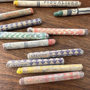 May include: A collection of vintage crayons in various colors, including yellow, orange, green, and purple, with geometric patterns and text. Some crayons have the text "VISUALIZER YELLOW" and "CRESTON CRAYON CO."