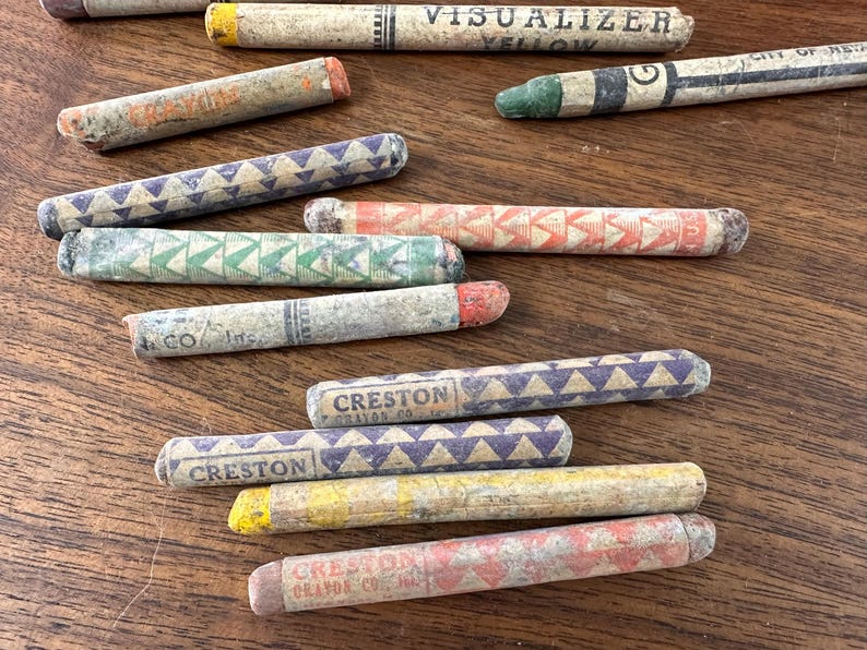 May include: A collection of vintage crayons in various colors, including yellow, orange, green, and purple, with geometric patterns and text. Some crayons have the text "VISUALIZER YELLOW" and "CRESTON CRAYON CO."