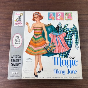 May include: A vintage Magic Mary Jane paper doll set box by Milton Bradley Company. The box features an illustration of a doll with red hair wearing a striped dress, surrounded by various outfits, including a green coat and a blue plaid suit.