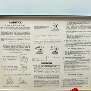 Puede incluir: Caja abierta del juego Slapstick, dise&ntilde;ado para 2, 3 o 4 jugadores. El interior presenta instrucciones y diagramas impresos sobre un fondo crema, con un borde rojo y azul. El t&iacute;tulo del juego es visible.