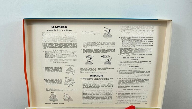 Puede incluir: Caja abierta del juego Slapstick, dise&ntilde;ado para 2, 3 o 4 jugadores. El interior presenta instrucciones y diagramas impresos sobre un fondo crema, con un borde rojo y azul. El t&iacute;tulo del juego es visible.