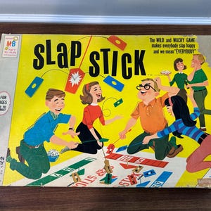 Puede incluir: Caja de juego de mesa vintage "Slap Stick" de Milton Bradley. La caja es amarilla y presenta ilustraciones de personas jugando. El texto en la caja incluye "Slap Stick", "Para edades de 10 a adulto" y "El juego salvaje y chiflado".