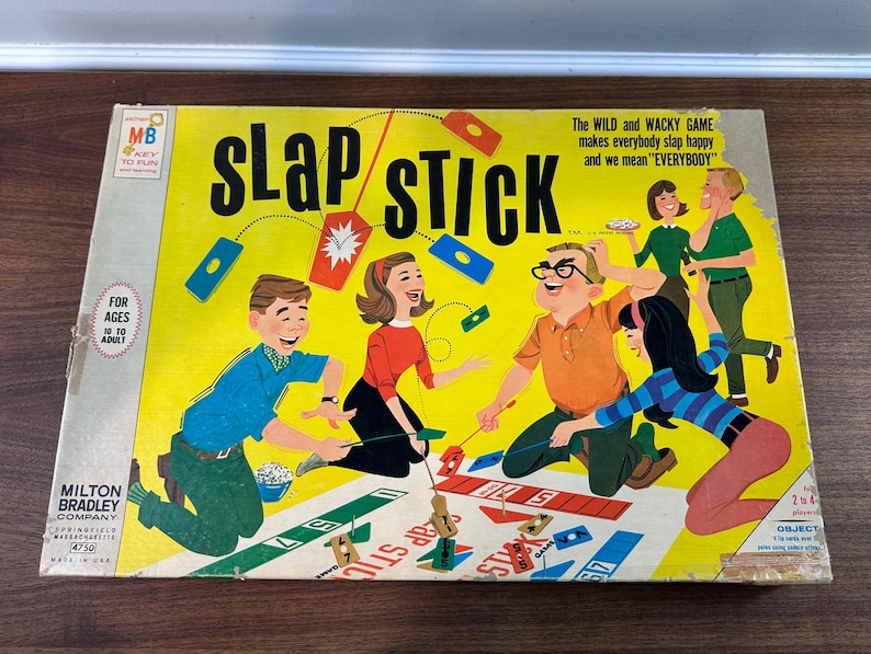 Puede incluir: Caja de juego de mesa vintage "Slap Stick" de Milton Bradley. La caja es amarilla y presenta ilustraciones de personas jugando. El texto en la caja incluye "Slap Stick", "Para edades de 10 a adulto" y "El juego salvaje y chiflado".