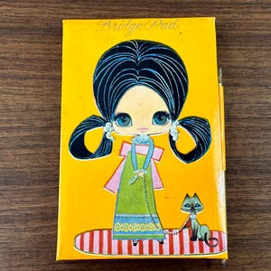 May include: A yellow vintage notepad with a cartoon girl with large eyes and a cat on a leash. The notepad has the text "Bridge Pad" on the front.