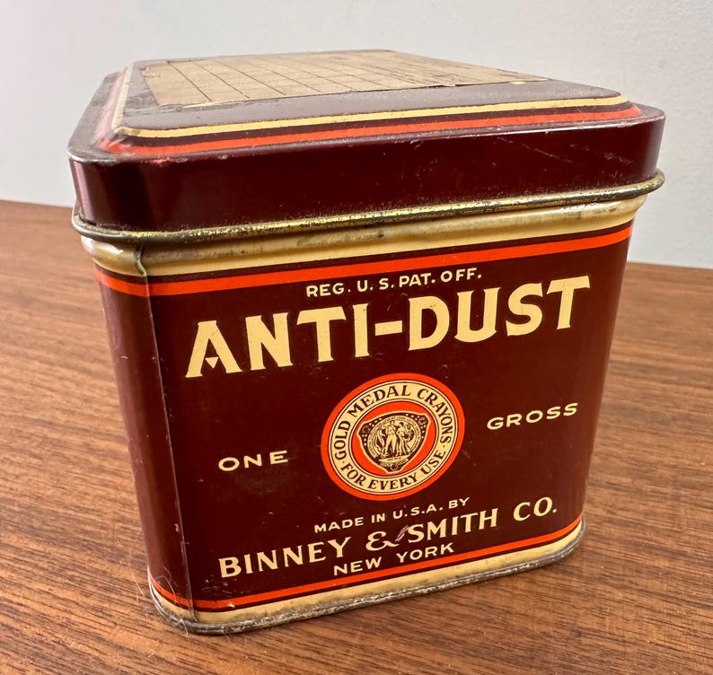 May include: A vintage, rectangular tin with a dark maroon body and gold accents. The tin reads "ANTI-DUST" in bold white letters, with "GOLD MEDAL CRAYONS" and "ONE GROSS" also visible. The bottom of the tin reads "MADE IN U.S.A. BY BINNEY & SMITH CO. NEW YORK."