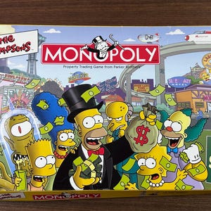 May include: A The Simpsons Monopoly board game box featuring a colorful illustration of Springfield characters including Homer, Marge, Bart, Lisa, Krusty the Clown, and Mr. Burns. The box displays the Monopoly logo, game title, and 6 collectible pewter tokens.