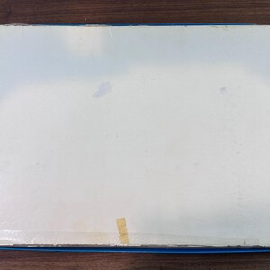 Puede incluir: Una caja rectangular con la parte superior blanca y bordes azules y rojos. La caja est&aacute; ligeramente desgastada, con algunas manchas y un trozo de cinta adhesiva. Descansa sobre una superficie de madera marr&oacute;n oscuro.
