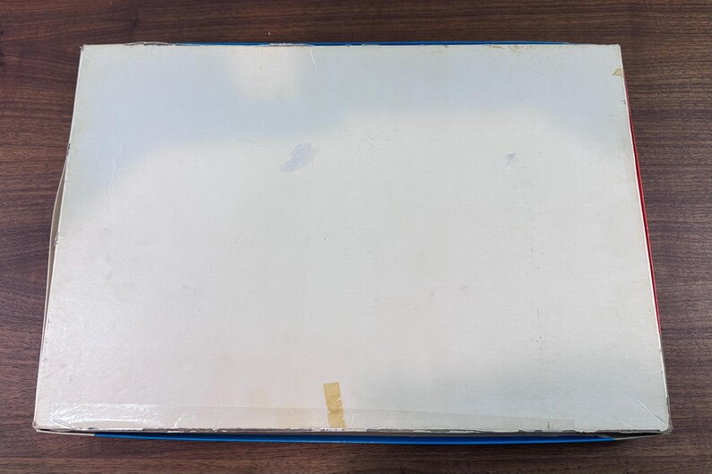 Puede incluir: Una caja rectangular con la parte superior blanca y bordes azules y rojos. La caja est&aacute; ligeramente desgastada, con algunas manchas y un trozo de cinta adhesiva. Descansa sobre una superficie de madera marr&oacute;n oscuro.