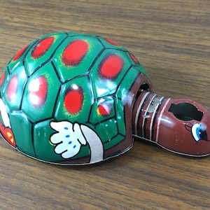 May include: A vintage wind-up toy turtle. The turtle's shell is green with red spots and a segmented pattern. The head and neck are brown with a smiling face and blue eyes. A white gloved hand is visible.