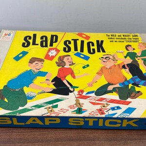 Puede incluir: Caja de juego de mesa vintage "Slap Stick" con un fondo amarillo e ilustraciones coloridas de personas jugando. La caja presenta el t&iacute;tulo del juego en letras grandes y el logotipo de Milton Bradley Company.
