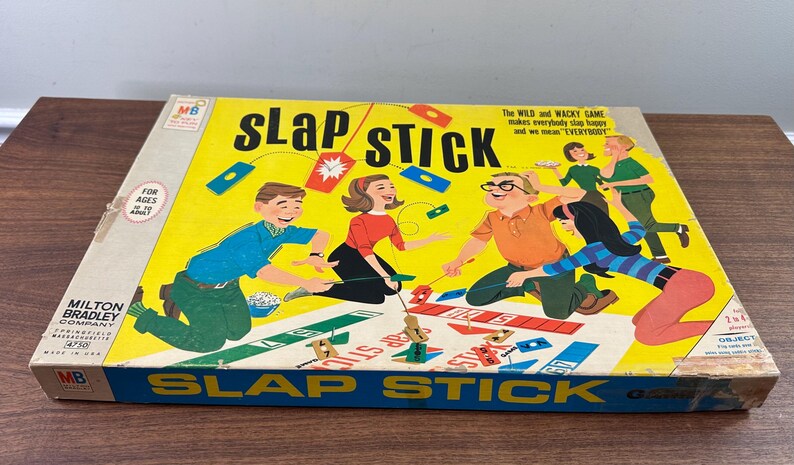 Puede incluir: Caja de juego de mesa vintage "Slap Stick" con un fondo amarillo e ilustraciones coloridas de personas jugando. La caja presenta el t&iacute;tulo del juego en letras grandes y el logotipo de Milton Bradley Company.