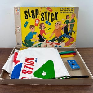 Puede incluir: Caja de juego vintage "Slap Stick" con fondo amarillo e ilustraciones de dibujos animados. La caja contiene piezas de juego, incluyendo palos de colores, cartas y un tablero de juego blanco grande con formas rojas, azules y verdes. La caja dice "Slap Stick Game".