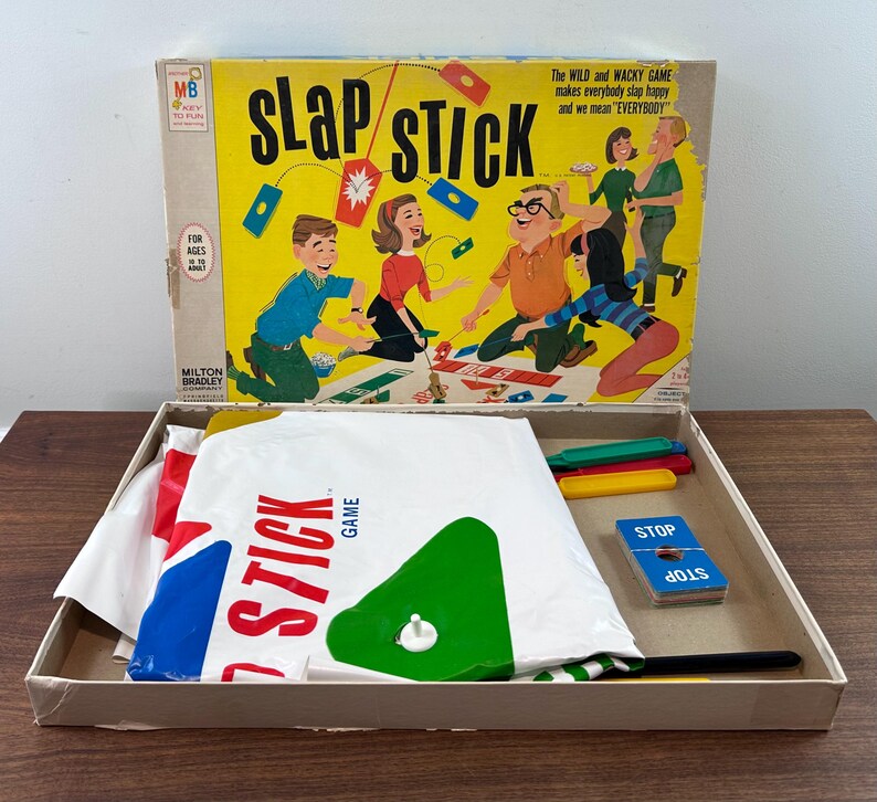 Puede incluir: Caja de juego vintage "Slap Stick" con fondo amarillo e ilustraciones de dibujos animados. La caja contiene piezas de juego, incluyendo palos de colores, cartas y un tablero de juego blanco grande con formas rojas, azules y verdes. La caja dice "Slap Stick Game".