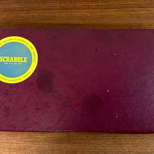 May include: A burgundy box with a yellow circle sticker that says "SCRABBLE REG. U.S. PAT. OFF. FOR PRODUCTION & MARKETING COMPANY MANUFACTURED BY SELCHOW & RIGHTER CO. NEW YORK, N.Y."