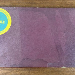 May include: A burgundy board game box with a yellow circle on the front. The circle has the word "SCRABBLE" in white letters and the text "Manufactured by Selchow & Righter Co. New York, N.Y." in black letters. The text "For Production & Marketing to" is also in black letters.