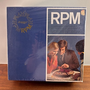 May include: A blue box with a game called RPM, a word game for quick thinking players. The box features a photo of three people playing the game. The text on the box reads "The rewarding word game for quick-thinking players. Letter tiles are placed on a revolving playing board and the more you make and fast turning, try and beat each other.  Digressing spelling and fast turning, try and beat each other."
