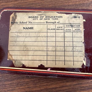 May include: A rectangular, maroon-colored metal box with a paper label. The label, from the Board of Education, City of New York, has handwritten entries under "Name" and "Class." The box has a red and white trim.