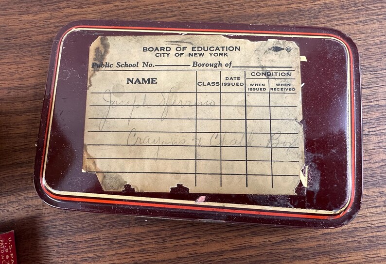 May include: A rectangular, maroon-colored metal box with a paper label. The label, from the Board of Education, City of New York, has handwritten entries under "Name" and "Class." The box has a red and white trim.