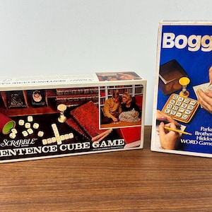 May include: Two board games, one is a Scrabble Sentence Cube Game with a brown wooden box and white dice, the other is a blue Boggle game with a white box and a wooden letter cube tray.