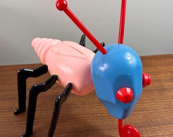 Puede incluir: Un insecto de juguete de plástico rosa y azul con patas negras y antenas rojas. El insecto tiene una cola en espiral y una cabeza grande con ojos rojos.