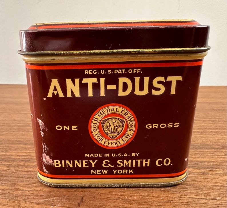May include: A vintage rectangular tin with a maroon and gold color scheme. The text "ANTI-DUST" is prominently displayed in large, gold letters. The tin also features the text "ONE GROSS" and "GOLD MEDAL CRAYONS". The manufacturer's name, "BINNEY & SMITH CO., NEW YORK" is also visible.