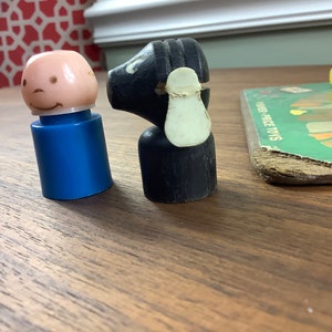 K&ouml;nnte beinhalten: Ein vintage Holzspielzeug schwarzes Schaf mit wei&szlig;em Gesicht und eine blaue Plastikspielzeugperson mit rosa Gesicht und einem L&auml;cheln. Die Spielzeuge befinden sich auf einer Holzoberfl&auml;che.