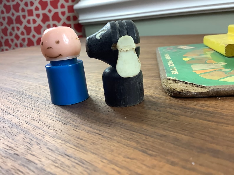 K&ouml;nnte beinhalten: Ein vintage Holzspielzeug schwarzes Schaf mit wei&szlig;em Gesicht und eine blaue Plastikspielzeugperson mit rosa Gesicht und einem L&auml;cheln. Die Spielzeuge befinden sich auf einer Holzoberfl&auml;che.