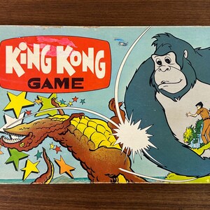 May include: Vintage King Kong board game box featuring a cartoon illustration of King Kong, a dinosaur-like creature, and people. The box has a red and white title, with text indicating it's for ages 5 to 12. Made by Milton Bradley.