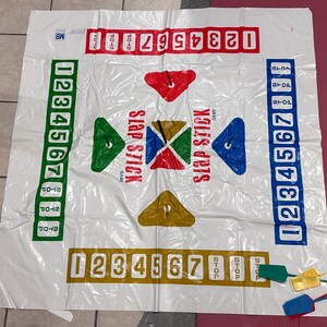 Puede incluir: Un tapete de juego Slap Stick de pl&aacute;stico blanco con gr&aacute;ficos y n&uacute;meros coloridos. El tapete presenta columnas numeradas, indicadores de parada y formas geom&eacute;tricas en rojo, verde, azul y amarillo. Incluye paletas de colores.