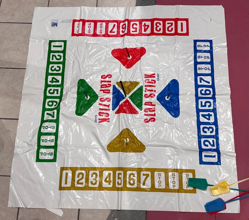 Puede incluir: Un tapete de juego Slap Stick de pl&aacute;stico blanco con gr&aacute;ficos y n&uacute;meros coloridos. El tapete presenta columnas numeradas, indicadores de parada y formas geom&eacute;tricas en rojo, verde, azul y amarillo. Incluye paletas de colores.