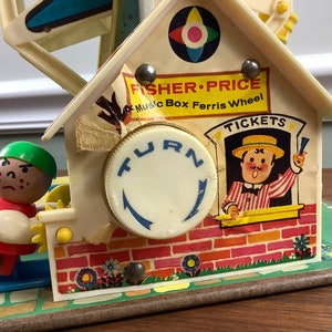 K&ouml;nnte beinhalten: Vintage Fisher-Price Musikbox-Riesenrad Spielzeug mit einer rot-wei&szlig;en Figur, einem Ticketschalter mit einem l&auml;chelnden Mann und einem Drehknopf. Das Spielzeug ist aus Kunststoff und hat ein farbenfrohes Design.