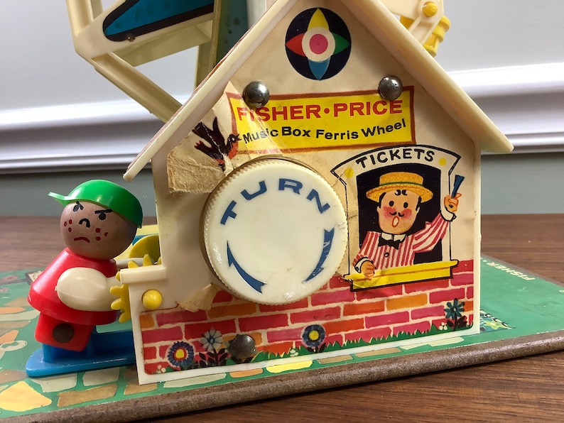 K&ouml;nnte beinhalten: Vintage Fisher-Price Musikbox-Riesenrad Spielzeug mit einer rot-wei&szlig;en Figur, einem Ticketschalter mit einem l&auml;chelnden Mann und einem Drehknopf. Das Spielzeug ist aus Kunststoff und hat ein farbenfrohes Design.