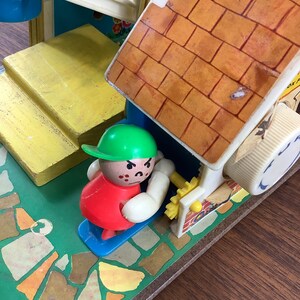 K&ouml;nnte beinhalten: Ein Vintage-Spielset mit einer kleinen, rot gekleideten Figur mit gr&uuml;nem Hut und einer wei&szlig;en Wurst. Die Figur steht auf einer blauen Plattform vor einem gelben und braunen Backsteinhaus mit wei&szlig;em Dach. Das Haus hat eine kleine, wei&szlig;e Kurbel an der Seite. Das Spielset steht auf einer gr&uuml;n-braunen gemusterten Oberfl&auml;che.