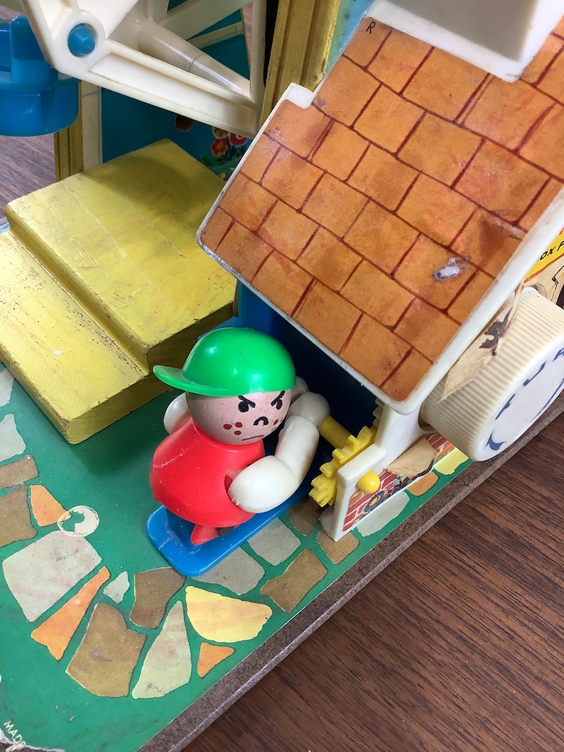 K&ouml;nnte beinhalten: Ein Vintage-Spielset mit einer kleinen, rot gekleideten Figur mit gr&uuml;nem Hut und einer wei&szlig;en Wurst. Die Figur steht auf einer blauen Plattform vor einem gelben und braunen Backsteinhaus mit wei&szlig;em Dach. Das Haus hat eine kleine, wei&szlig;e Kurbel an der Seite. Das Spielset steht auf einer gr&uuml;n-braunen gemusterten Oberfl&auml;che.