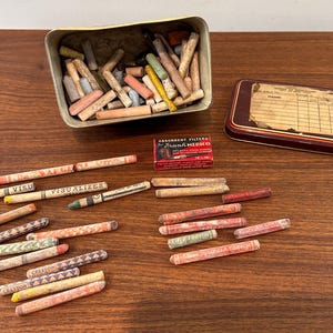 May include: An assortment of vintage crayons in various colors, scattered on a wooden surface. A metal tin and a small box of absorbent filters are also present. Some crayons are labeled with the words "Crayon" and "Visualizer".